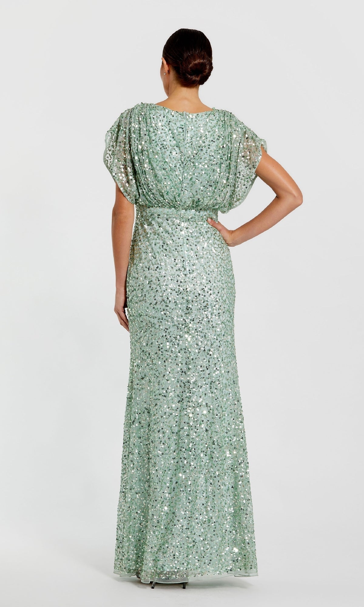 A woman faces away, wearing the Mac Duggal 5640 Short Sleeve Long Sequin Formal Dress—a floor-length, light green gown with sequins, short sleeves, and a belted waist.
